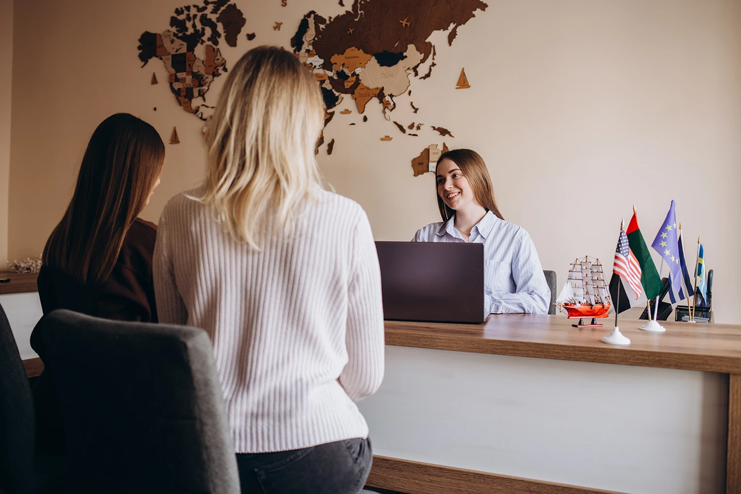 Consulente di viaggio sorride mentre ascolta due clienti al banco, mappa del mondo alle spalle