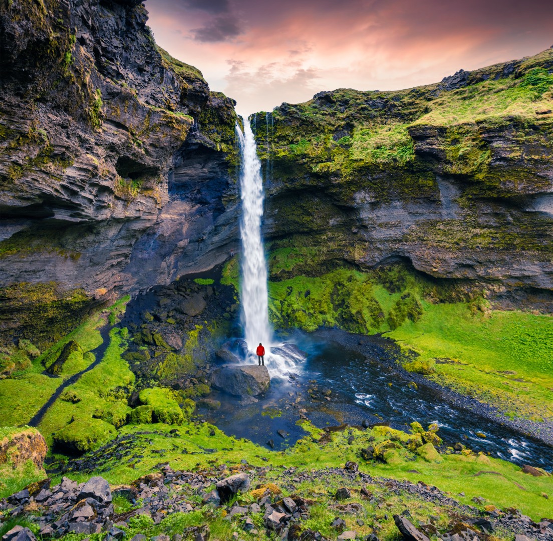 Viaggiatore in giacca rossa ai piedi di una cascata islandese tra le rocce verdi